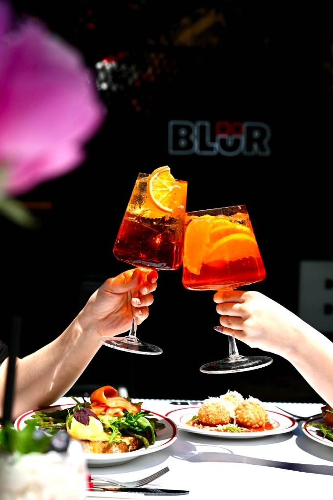 Două pahare de spritz ciocnite în fața logo-ului BLÜR, noaptea